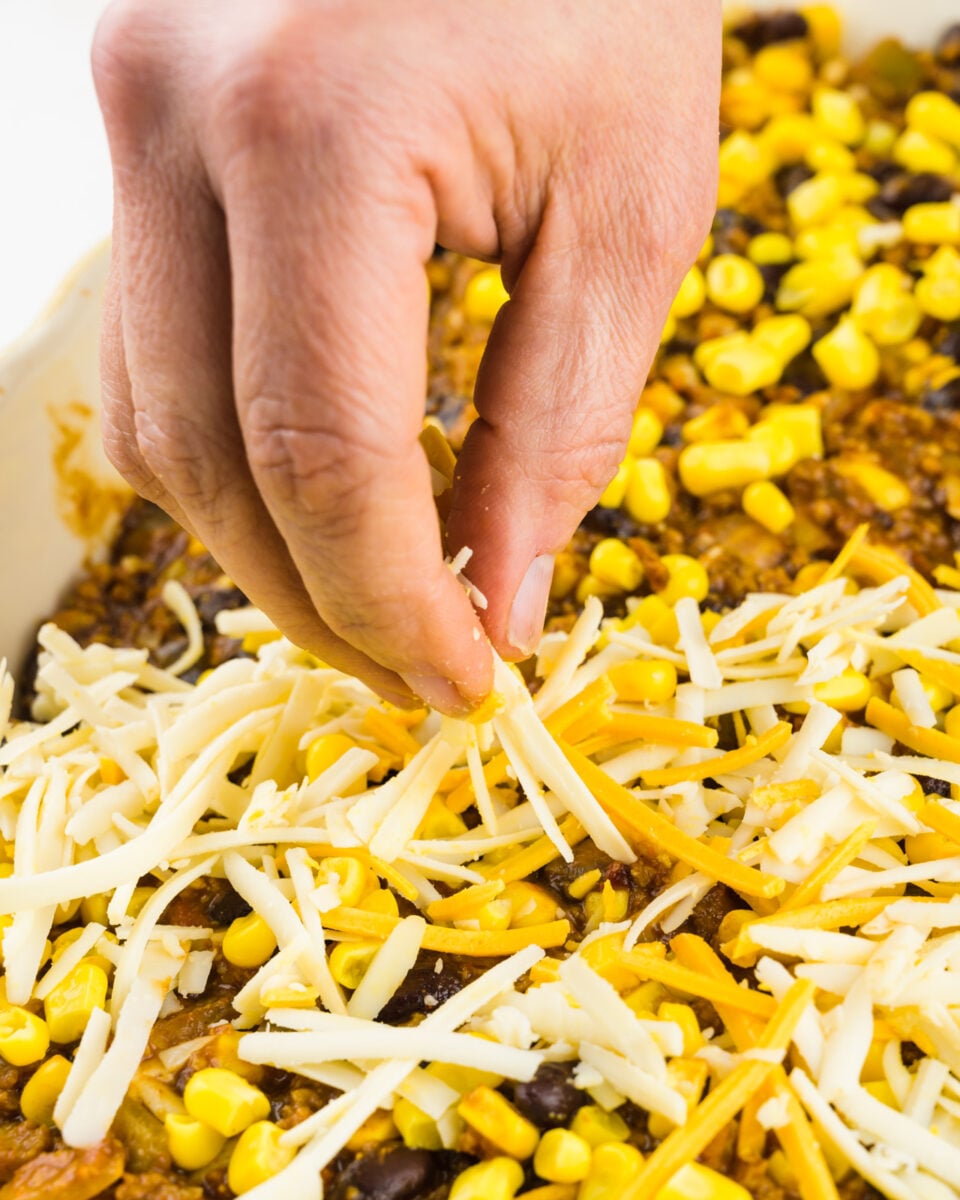 A hand holds vegan cheese and is distributing it over the top of a casserole.