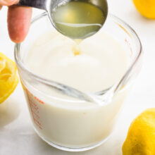 A hand holds a measuring spoon and is pouring lemon juice into plant-based milk in a glass measuring cup.
