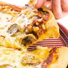 A hand holds a piece of pizza, pulling it out from a pan full of more pizza.