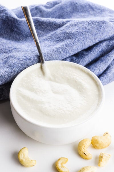 A white bowl holds cashew cream. There's a blue towel behind it and some cashews in front of it.