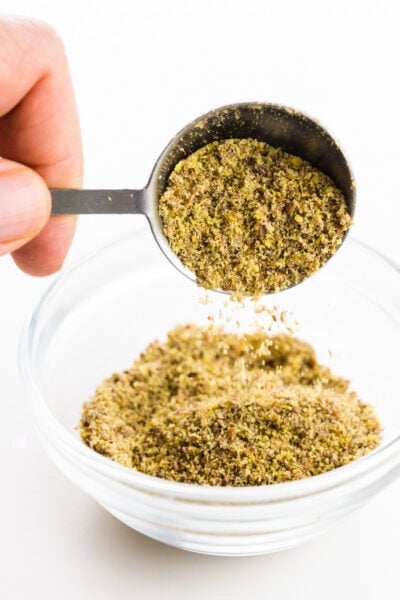 A hand holds a measuring spoon, pouring ground flax seeds into a small bowl.