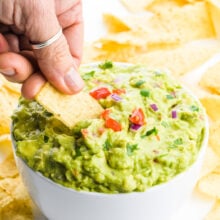 A hand holds a tortilla chip dipping it into a bowl of guacamole. There are tortilla chips behind it.