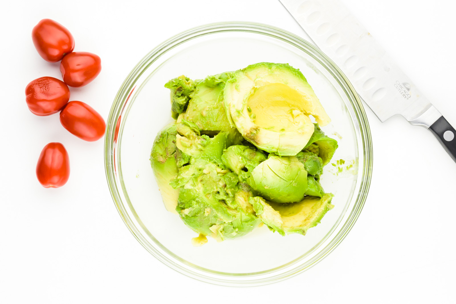 Looking down on a bowl of peeled avocados. There are cherry tomatoes beside it and a knife.