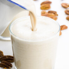 A pitcher pours a creamy milk into a small glass. There are pecans around it.
