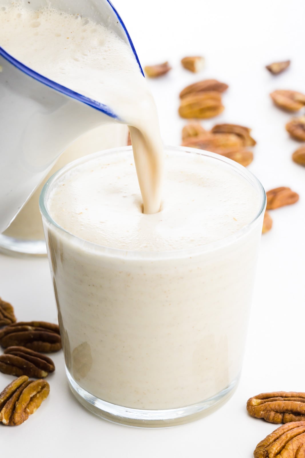 A pitcher pours a creamy milk into a small glass. There are pecans around it.