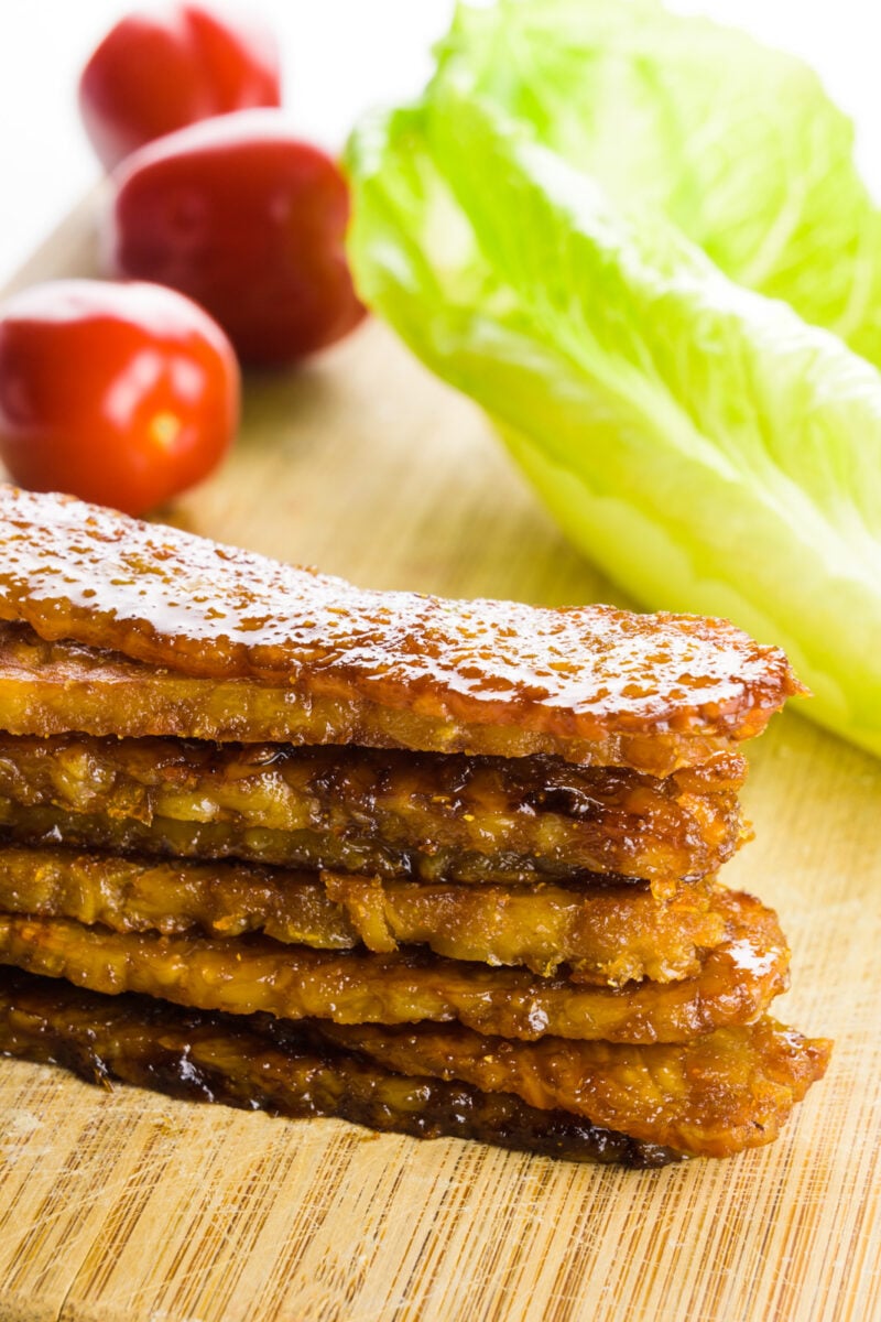 Several strips of tempeh bacon are stacked on a cutting board. There are cherry tomatoes and pieces of lettuce behind it.