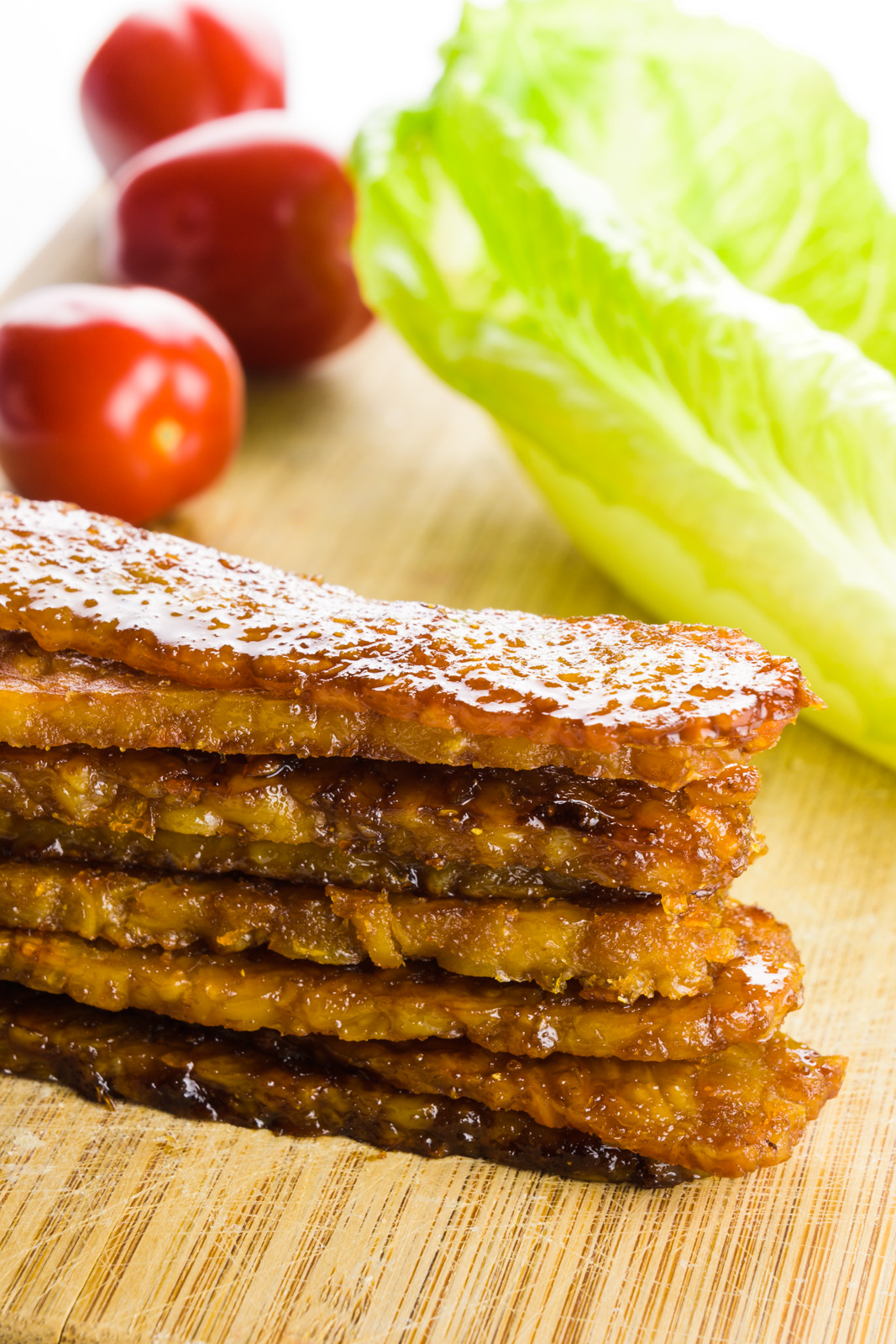 Several strips of tempeh bacon are stacked on a cutting board. There are cherry tomatoes and pieces of lettuce behind it.
