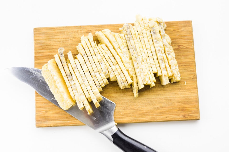 Thinly sliced tempeh sits on a cutting board next to a knife.