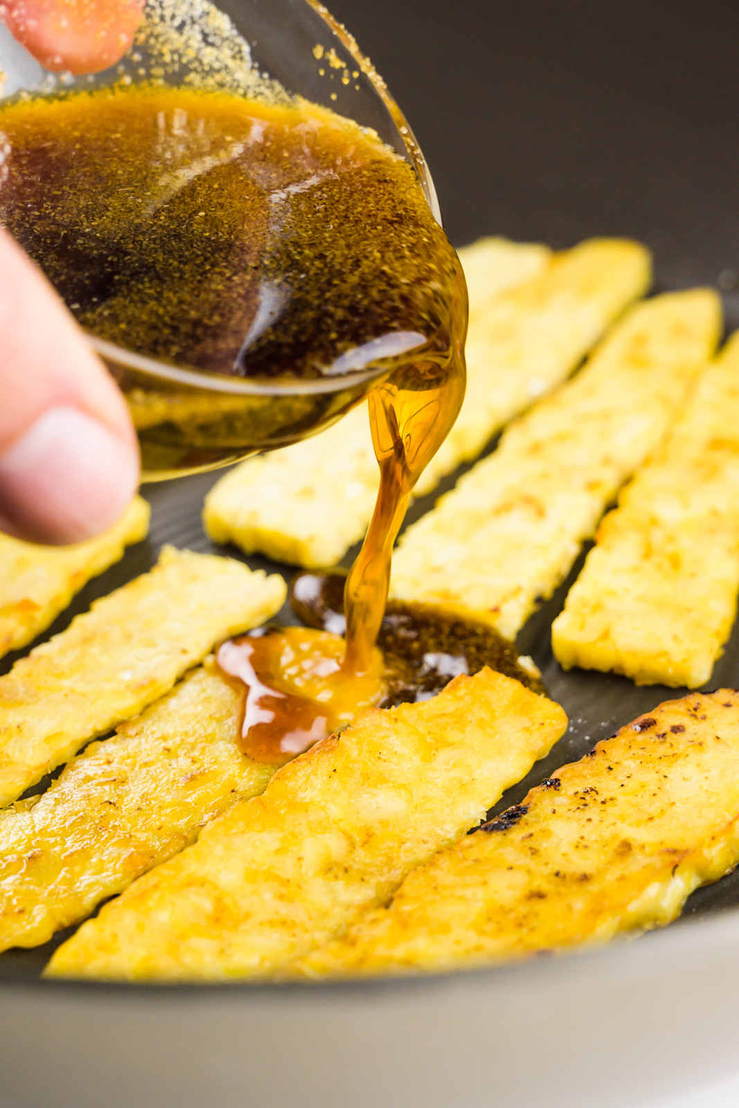 A hand holds a bowl of marinade and is pouring it over tempeh strips in a skillet.