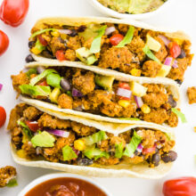 Looking down on 3 soft tacos with cherry tomatoes around it. There's also a bowl of salsa and a bowl of guacamole by it.