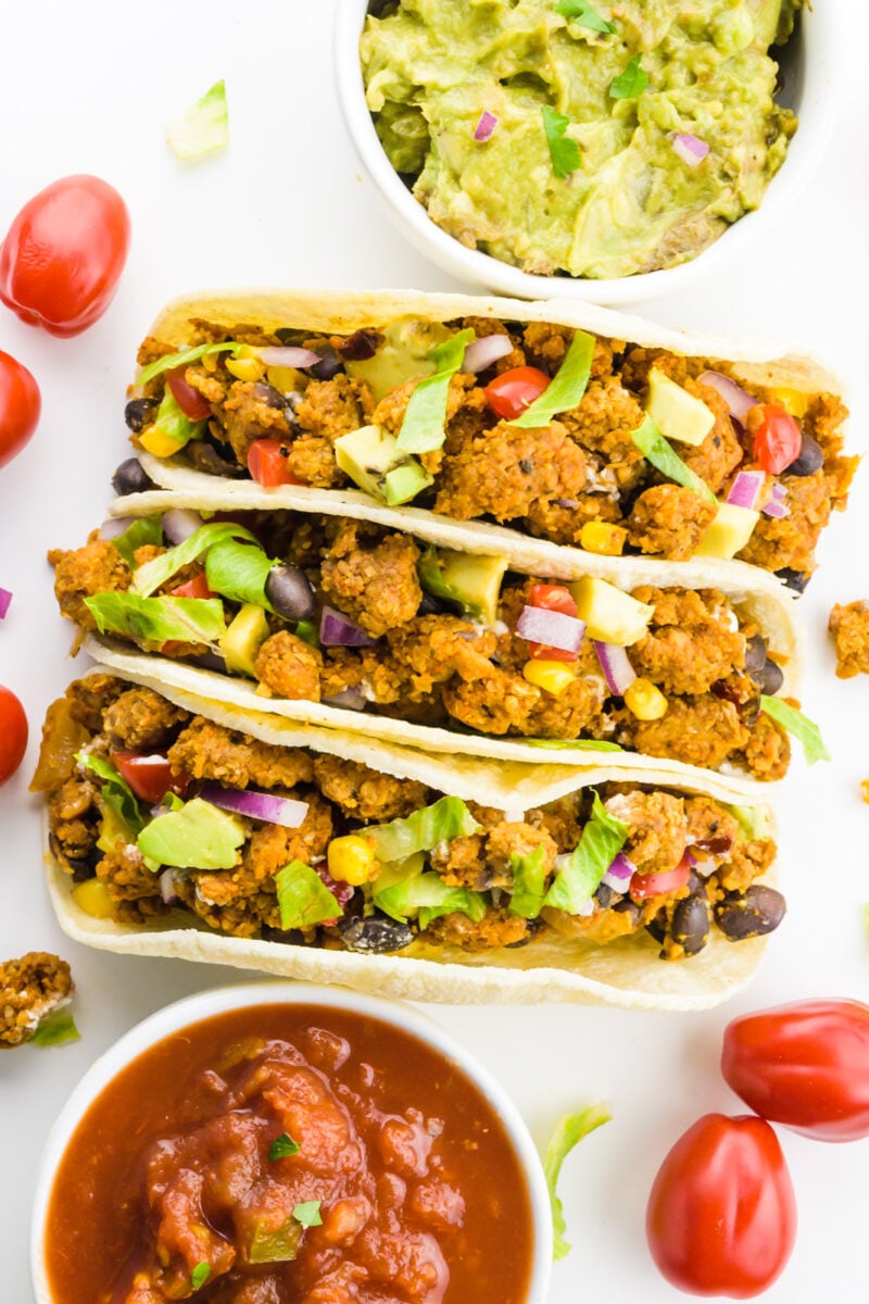 Looking down on 3 soft tacos with cherry tomatoes around it. There's also a bowl of salsa and a bowl of guacamole by it.