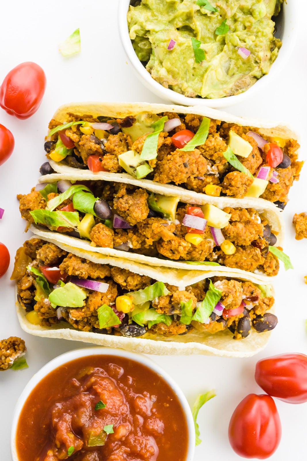 Looking down on 3 soft tacos with cherry tomatoes around it. There's also a bowl of salsa and a bowl of guacamole by it.