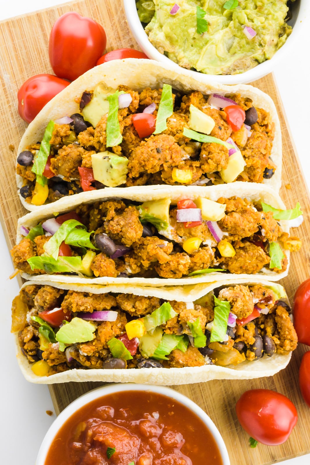 A closeup of three vegan tacos on a cutting board with cherry tomatoes and bowls of guacamole and salsa around it.