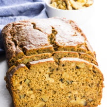 Looking straight on at a lot of bread with slices cut out. There's a blue kitchen towel and a bowl of walnuts behind it.