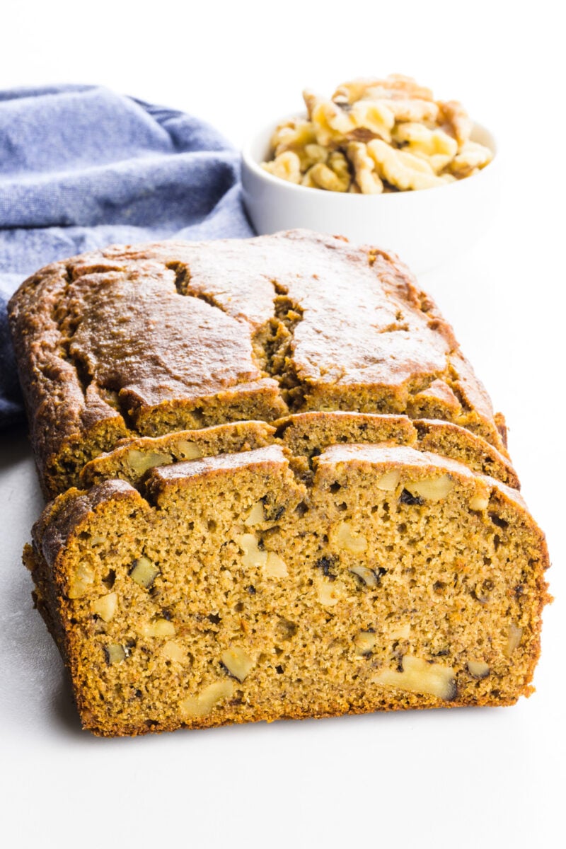 Looking straight on at a lot of bread with slices cut out. There's a blue kitchen towel and a bowl of walnuts behind it.