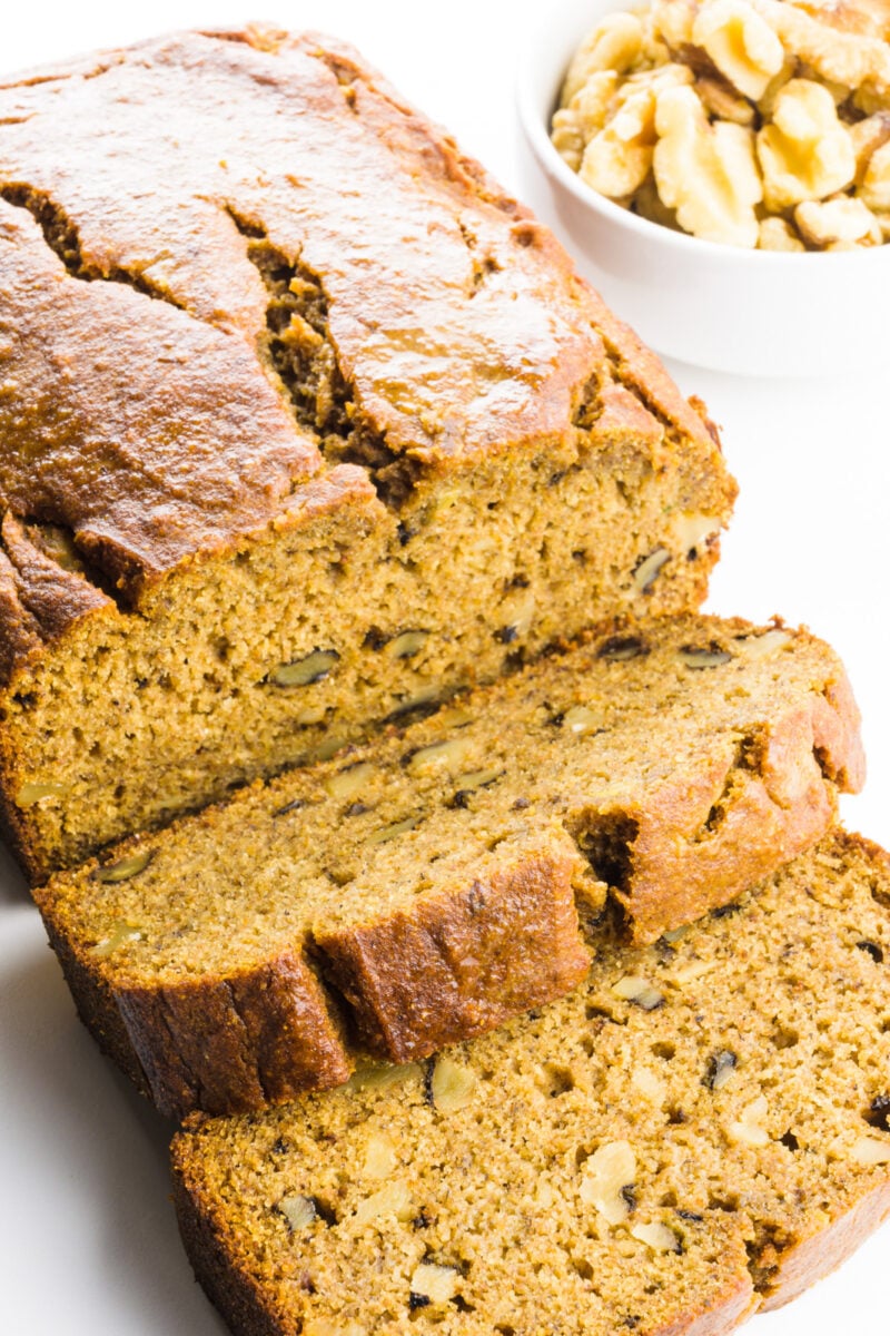 A loaf of whole wheat banana bread has several slices cut out. There's a small bowl of walnuts behind it.