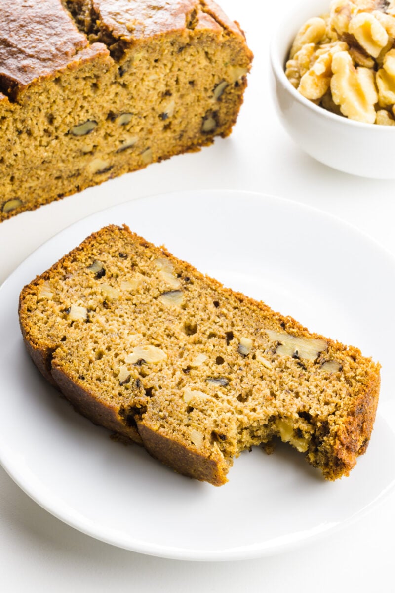 A slice of banana bread has a bite taken out of it and is sitting on a plate in front of the rest of the loaf and a bowl of walnuts.