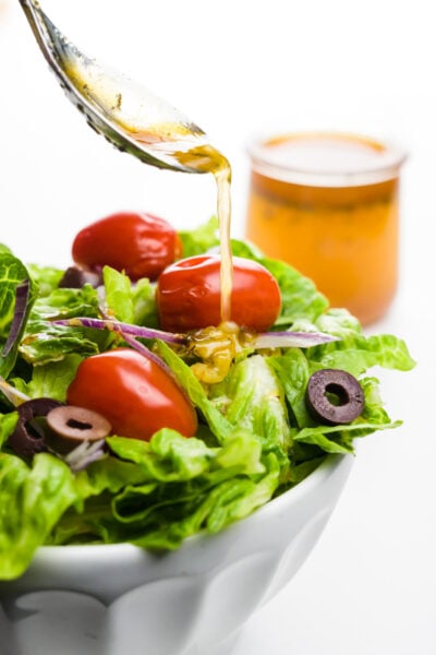 A spoon pours dressing over a salad. A jar with more dressing sits behind it.