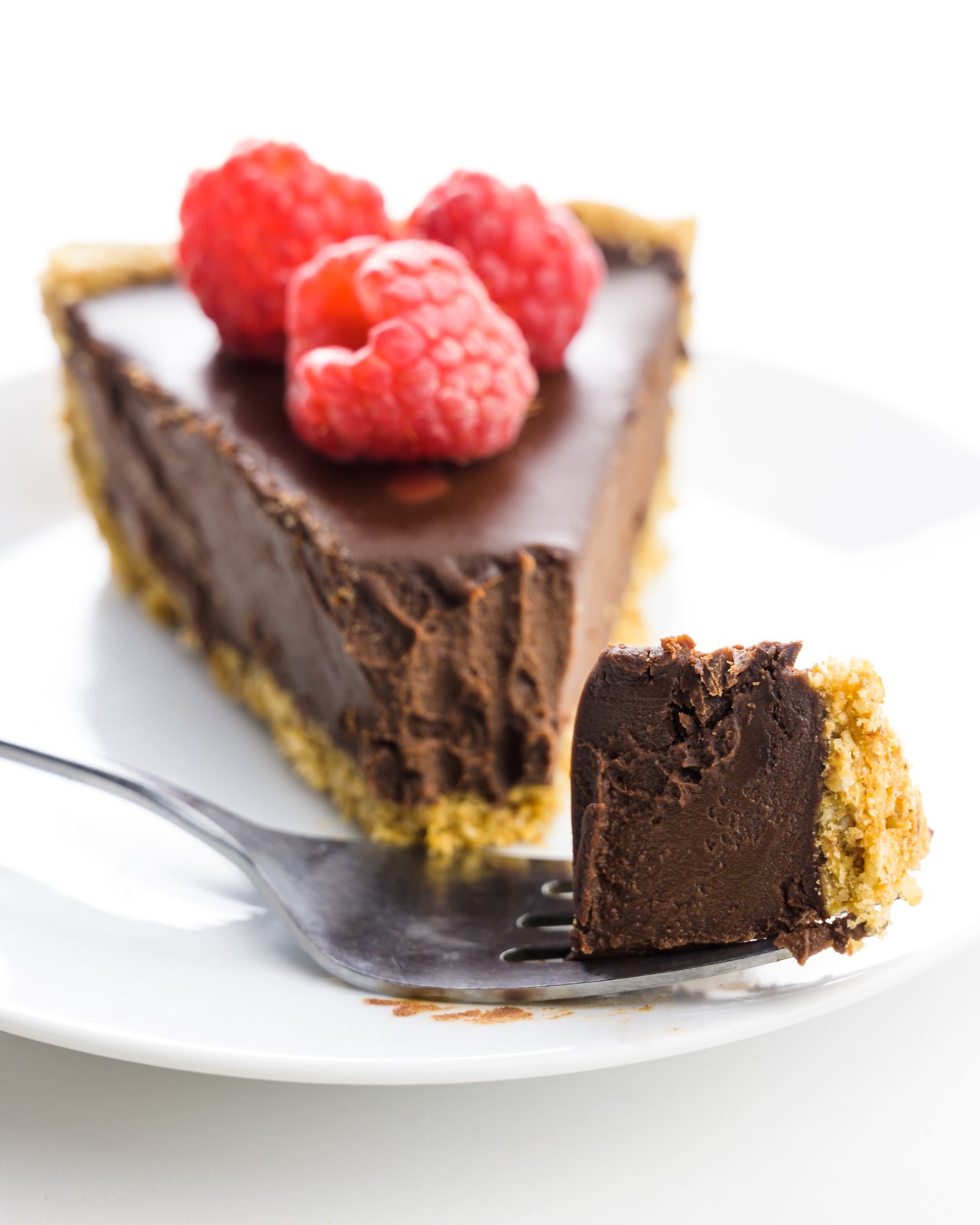 A fork holds a bite of chocolate pie sitting on a plate in front of the rest of the slice.. The slice is topped with fresh raspberries.