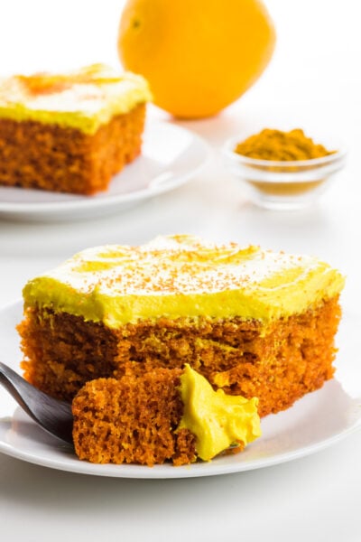 A slice of turmeric cake sits on a plate with a bite sitting on a fork. An orange and a small bowl of turmeric is behind it.