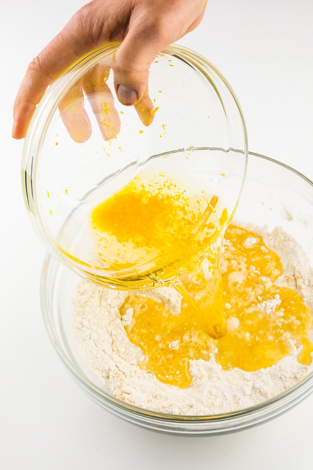 A hand holds the bowl of wet ingredients pouring it into the bowl with flour ingredients.