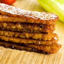 A stack of tempeh bacon sits on a cutting board in front of lettuce and a cherry tomato.