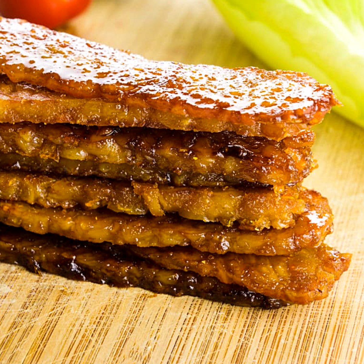 A stack of tempeh bacon sits on a cutting board in front of lettuce and a cherry tomato.