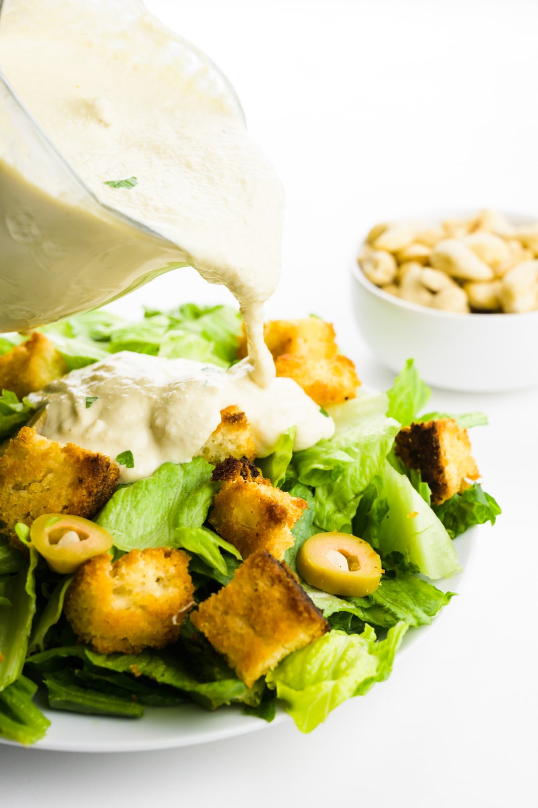 A serving dish full of vegan caesar dressing is being poured over a salad. There's a bowl of cashews in the background.
