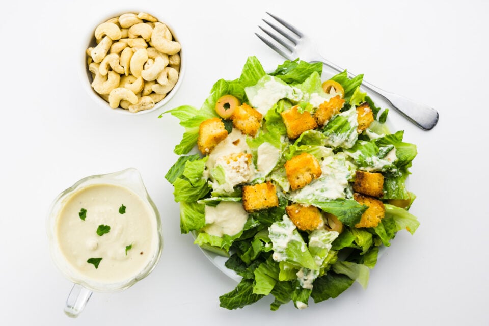 Looking down on a vegan caesar salad with a fork, dressing, and a bowl of cashews next to it.
