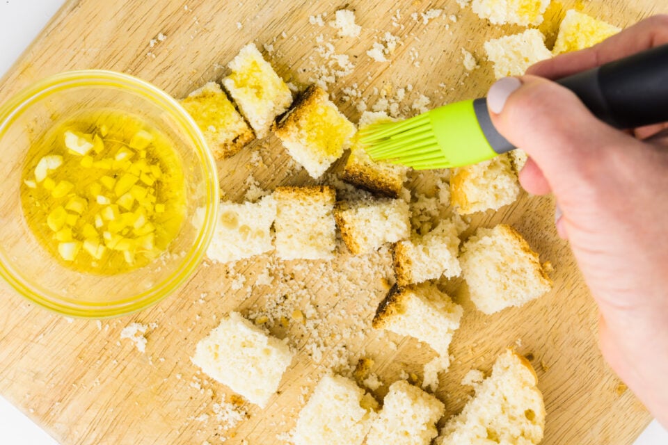 A hand holds a brush, dabbing oil over bread cubes to make croutons.