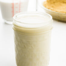 A small mason jar holds vegan evaporated milk. Behind it is a measuring glass with plant-based milk and a pie crust.