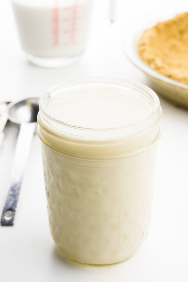Evaporated milk is in a glass jar. There's a measuring spoon beside it, a measuring cup and a pie crust behind it.