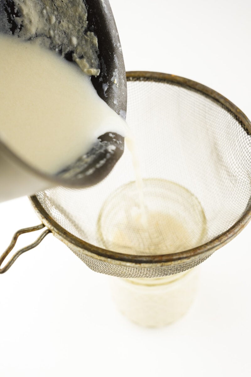 Vegan evaporated milk is being poured from a saucepan into a measuring cup.