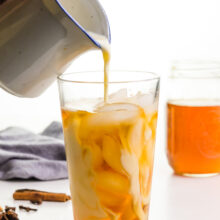 A hand holds a small pitcher, pouring plant milk into a glass with ice and tea in it. A jar is full of contracted tea behind it.