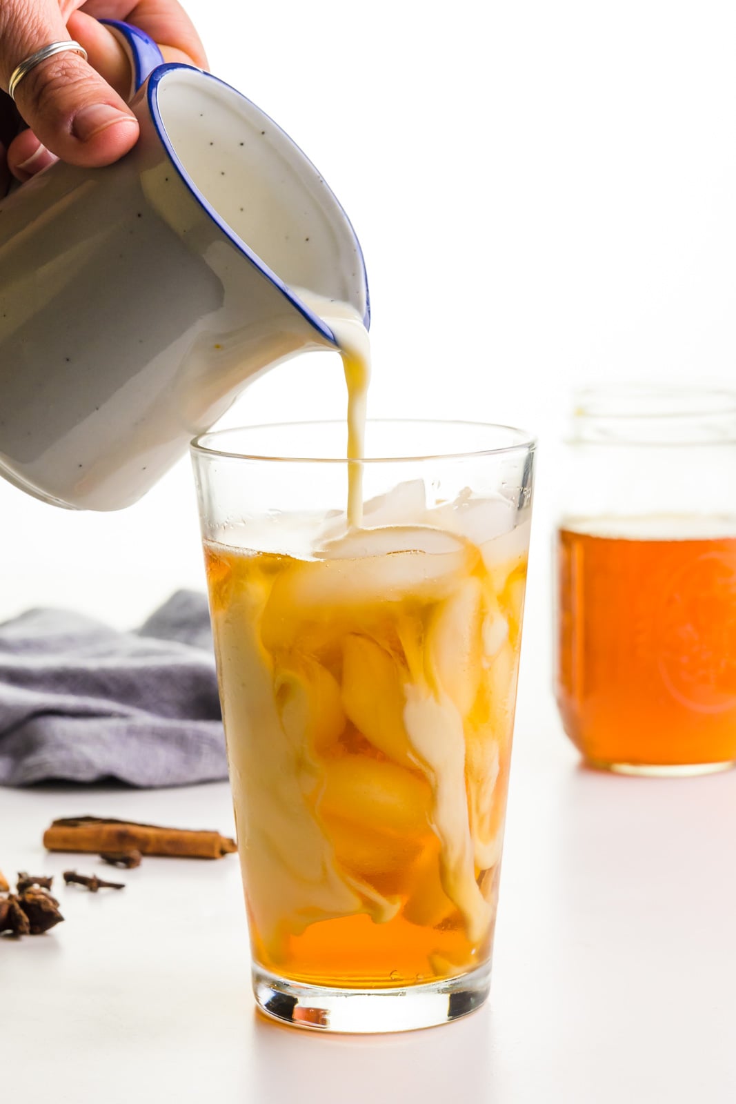 A hand holds a small pitcher, pouring plant milk into a glass with ice and tea in it. A jar is full of contracted tea behind it.