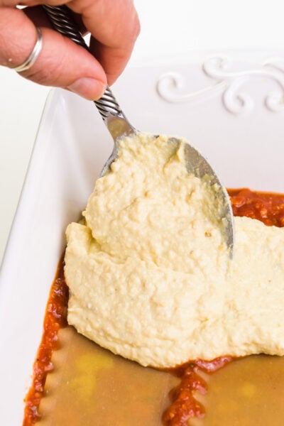 A hand holds a spoon, spreading vegan ricotta over lasagna noodles and sauce in a baking dish.