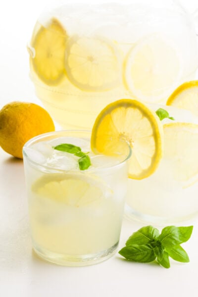 Two glasses of lemonade have fresh lemon slices on the glasses. There's a lemon behind it, and basil sprigs in front of it. There's a pitcher of more of the beverage behind it.