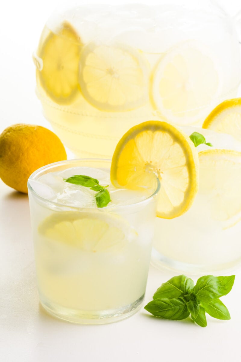 Two glasses of lemonade have fresh lemon slices on the glasses. There's a lemon behind it, and basil sprigs in front of it. There's a pitcher of more of the beverage behind it.