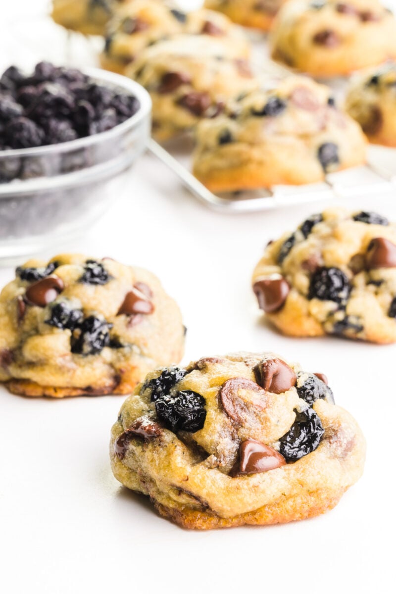 Several blueberry chocolate chip cookies are on a white counter. There's a wire wrack with more cookies cooling behind them and a bowl full of dried blueberries.