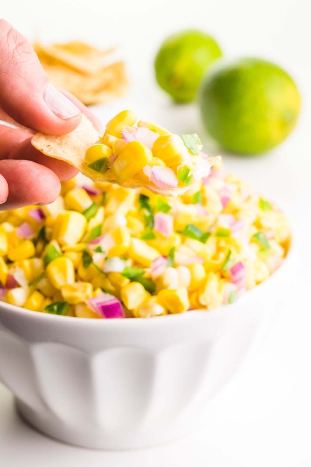 A hand holds a tortilla chip with corn salsa on it. It's sitting above a bowl full of the salsa. There are limes behind it.