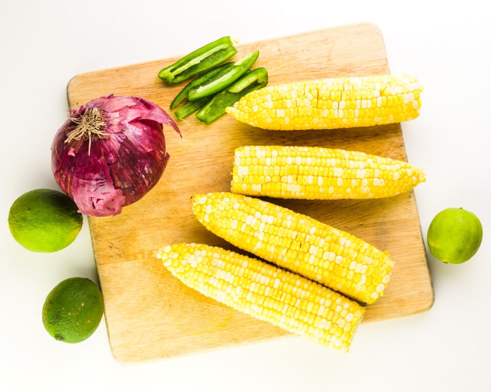 Looking down on a cutting board with 4 ears of corn, a red onion, chopped jalapeños, and several whole limes.