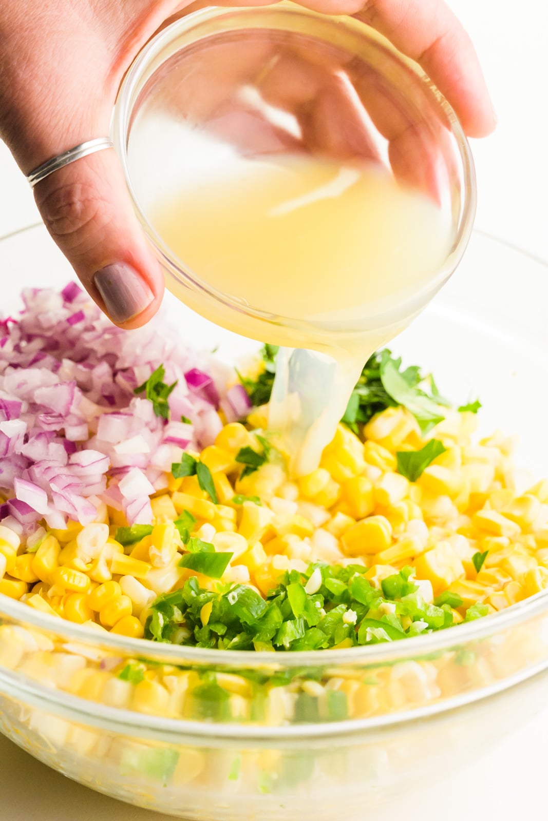 A hand holds a small bowl pouring lime juice over another bowl full of corn, red onions, and chopped jalapeños. 