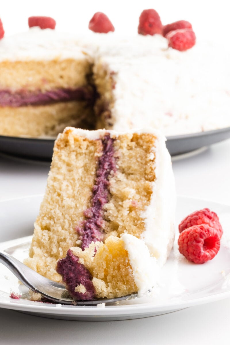 A slice of cake sits on its side on a plate. A fork has taken a bite out of it and this sits in front of the slice. The rest of the cake is behind it.