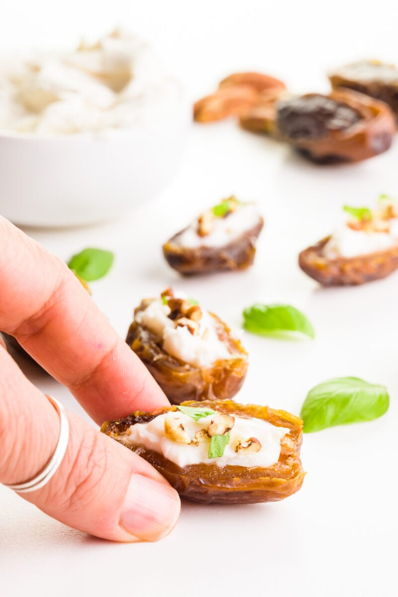 A hand reaches in and grabs a stuffed date. There are more dates with cream cheese filling behind it, along with basil leaves.