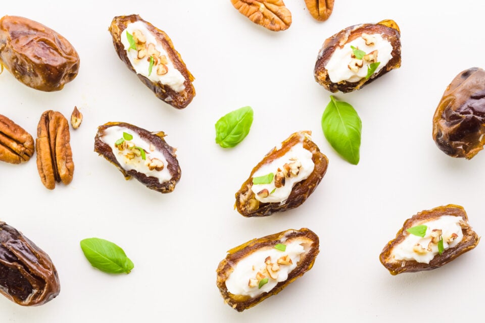 Looking down on a white counter with dates stuffed with cream cheese. There are pecans and green basil leaves between the dates.