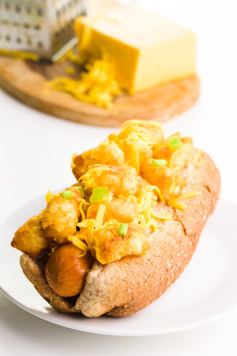 A hot dog is in a bun and is topped with tater tots and cheese. A cutting board is behind it with more cheese and a grater.
