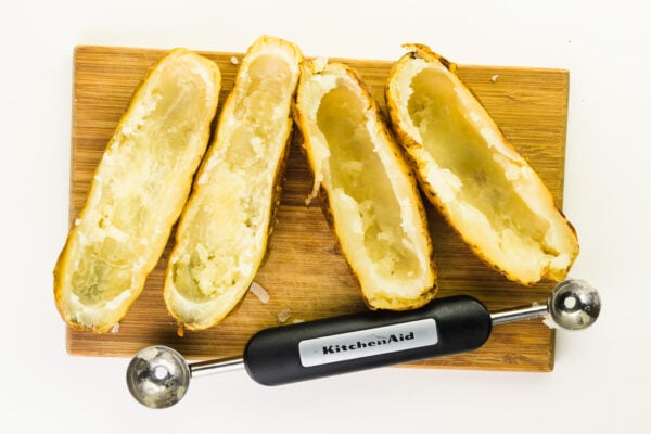 Four baked potatoes have had the centers scooped out. They're sitting on a cutting board next to a melon baller that was used to scoop the potatoes.