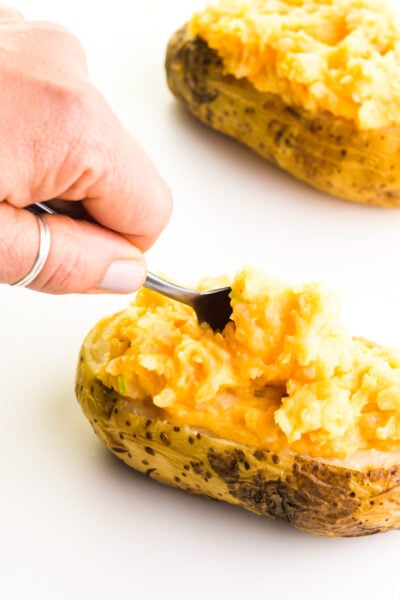 A hand holds a spoon and is placing cheesy mashed potato filling into a scooped out baked potato. Another potato sits behind it.