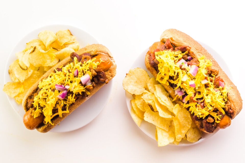 Looking down on two vegan hot dogs topped with chili and cheese. There are potato chips sitting by both.