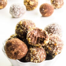 Several vegan truffles are in a bowl with more truffles behind them. They're all coated with different toppings, like chopped nuts and coconut.